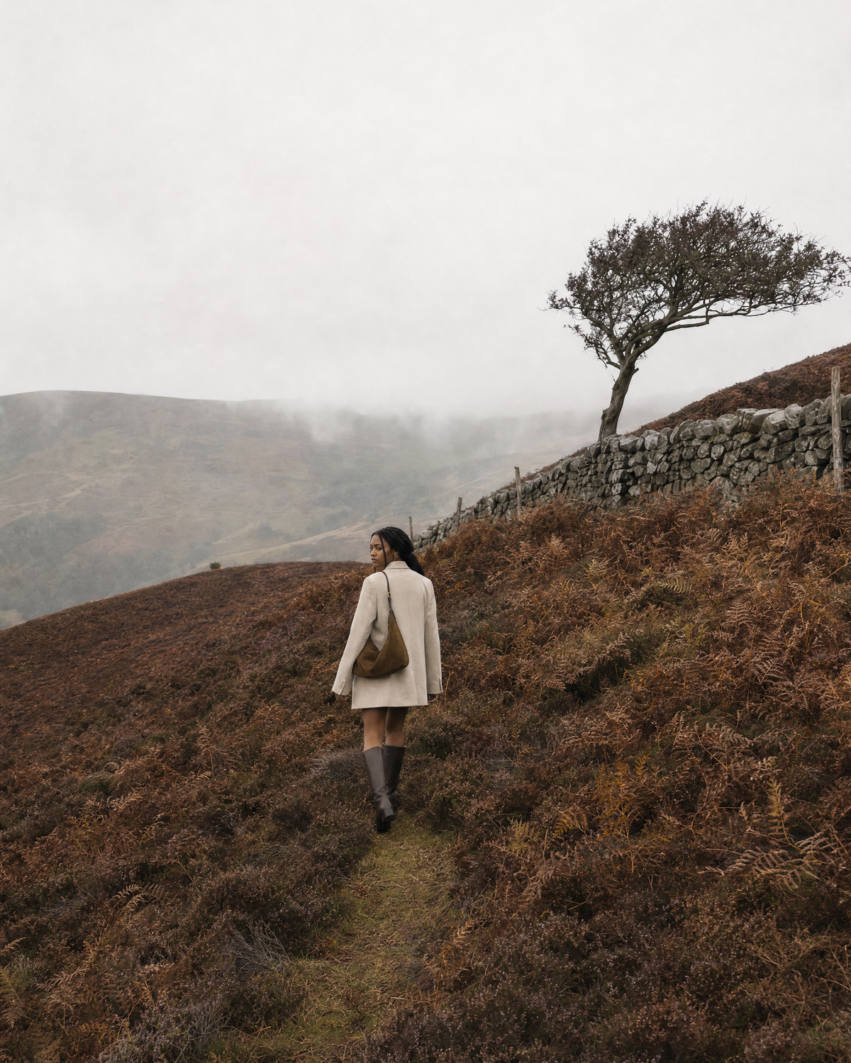 Woman walking along a dry-stone wall on a misty moor with The Brume Petite in Tabac