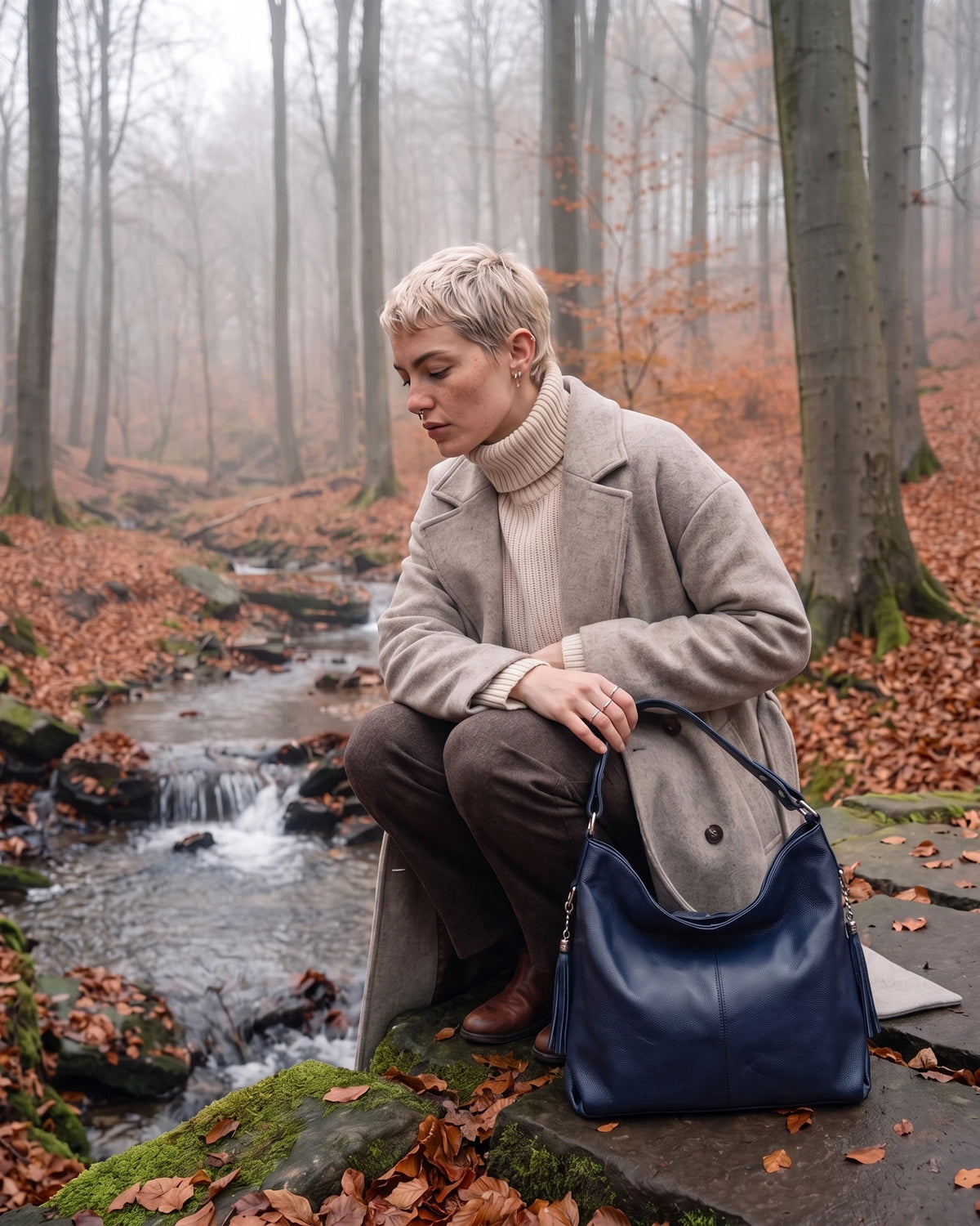 The Houle Soft in Nuit, midnight navy leather hobo bag on a stone beside a forest stream, model crouching