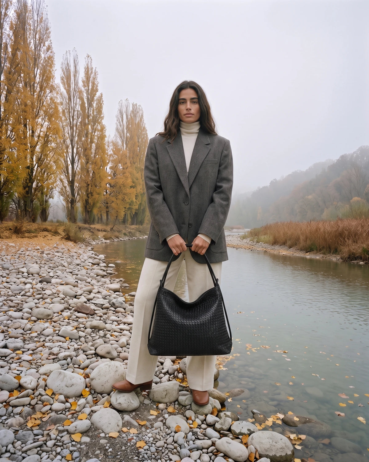 The Embellie Grand in Noir Mat, large matte black woven leather tote held in front of her by the river