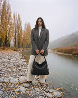The Embellie Grand in Noir Mat, large matte black woven leather tote held in front of her by the river