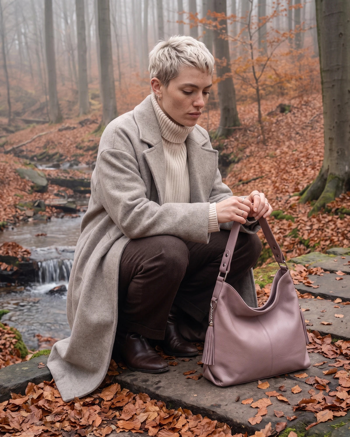 The Houle Soft in Rose, dusty rose leather hobo bag beside a model crouching by a stream in a misty autumn beech forest