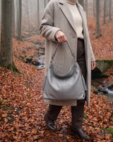 The Houle Soft in Galet, pebble grey leather hobo bag carried by the short handle in a misty autumn beech forest