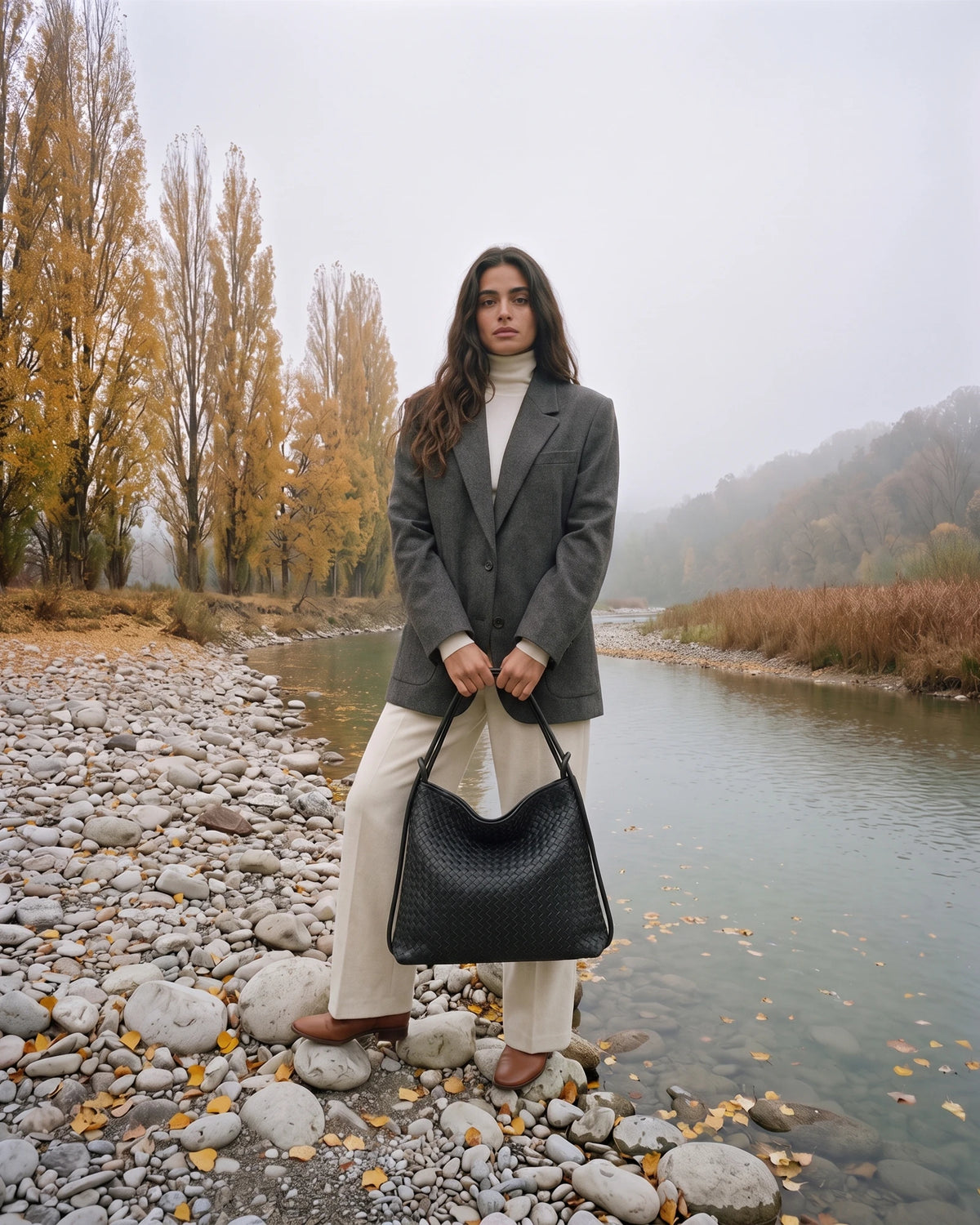 The Embellie in Noir Mat, matte black woven leather tote held in front of her by the river