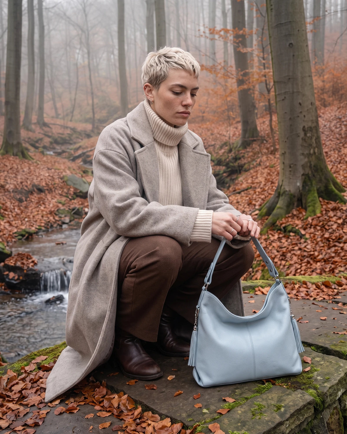 The Houle Soft in Ciel, powder blue leather hobo bag on a stone beside a forest stream, model crouching