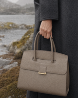 Hand holding the top handles of The Aurore Briefcase in Sable in the rain
