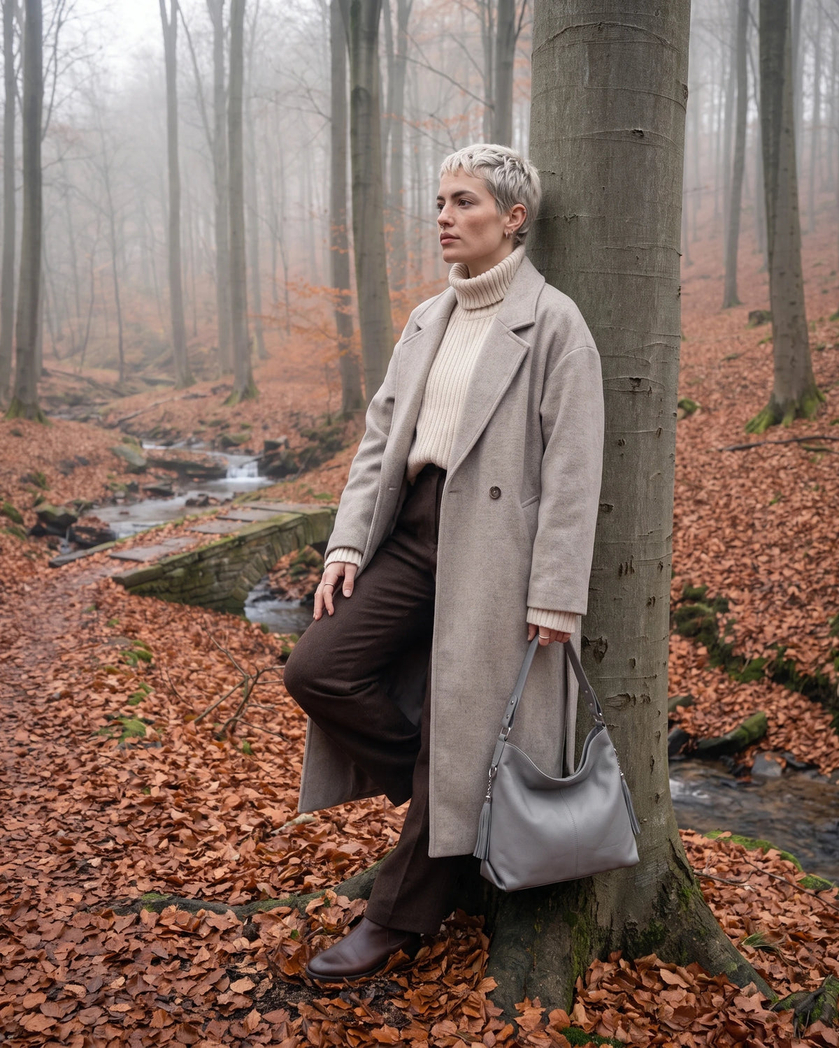 The Houle Soft in Galet, pebble grey leather hobo bag held by the short handle, model leaning against a beech tree in autumn mist