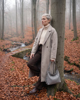 The Houle Soft in Galet, pebble grey leather hobo bag held by the short handle, model leaning against a beech tree in autumn mist