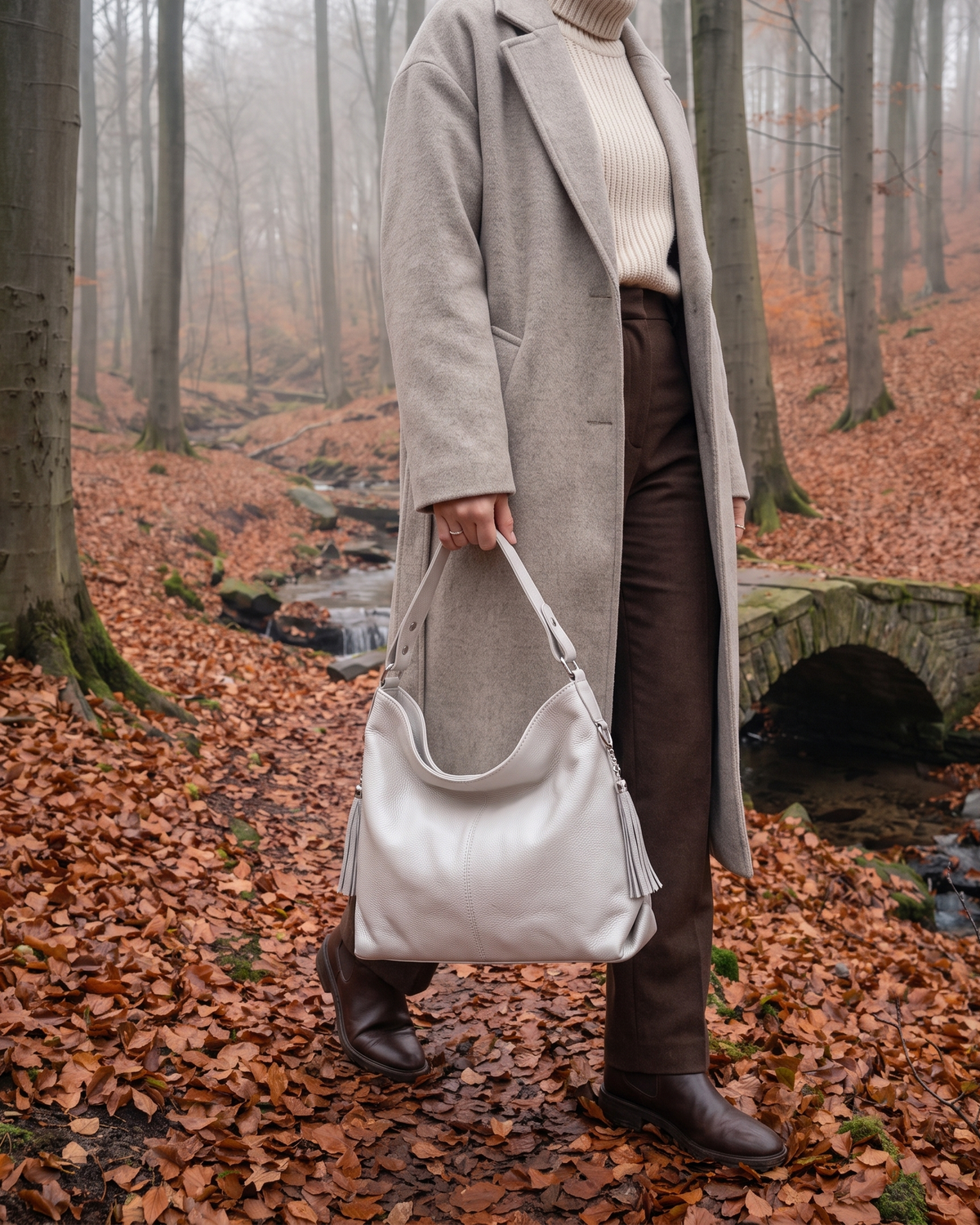 The Houle Soft in Écru, off-white leather hobo bag carried by the handle in a misty autumn beech forest