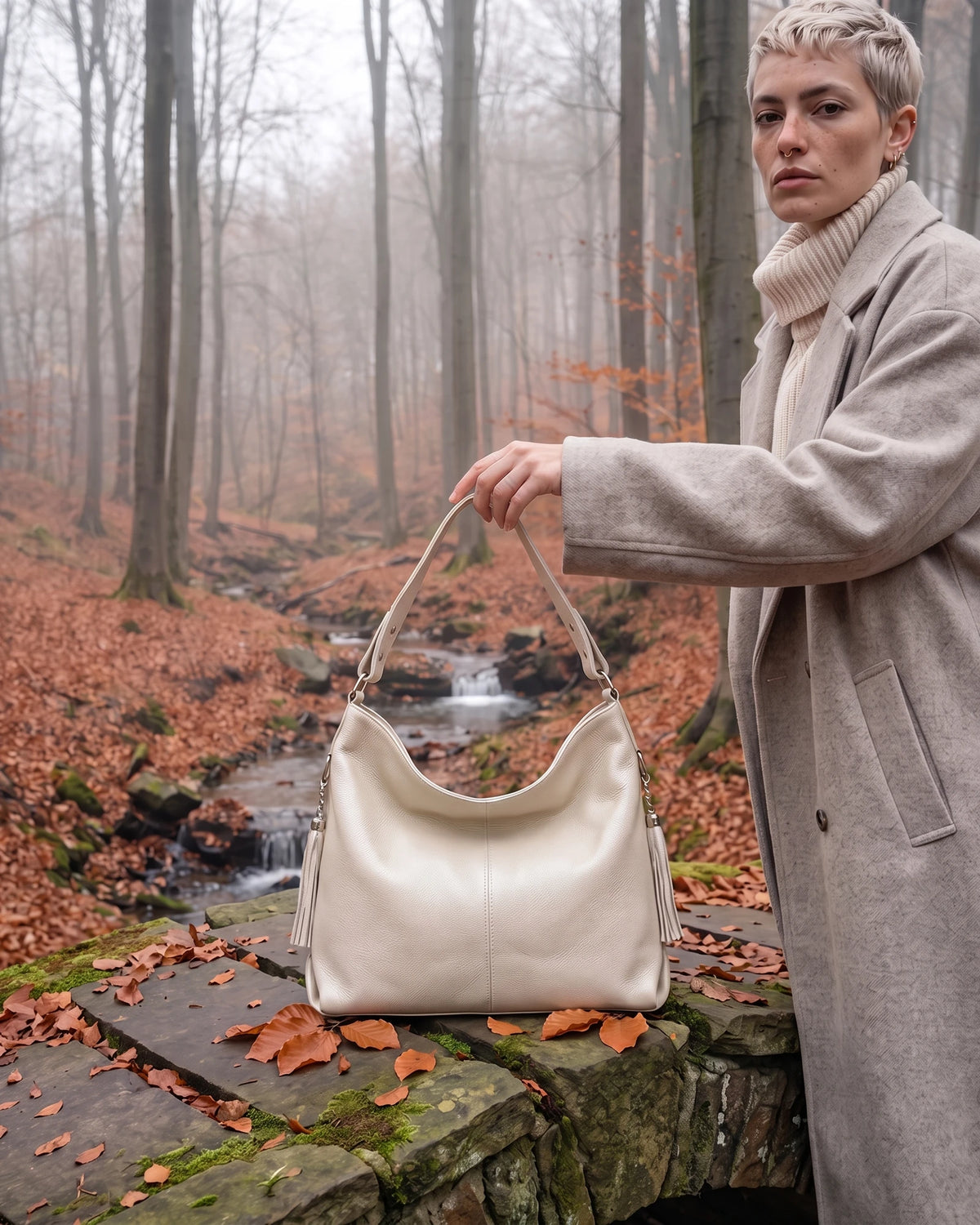 The Houle Soft in Sable, ivory sand leather hobo bag held above the mossy stones of a forest footbridge