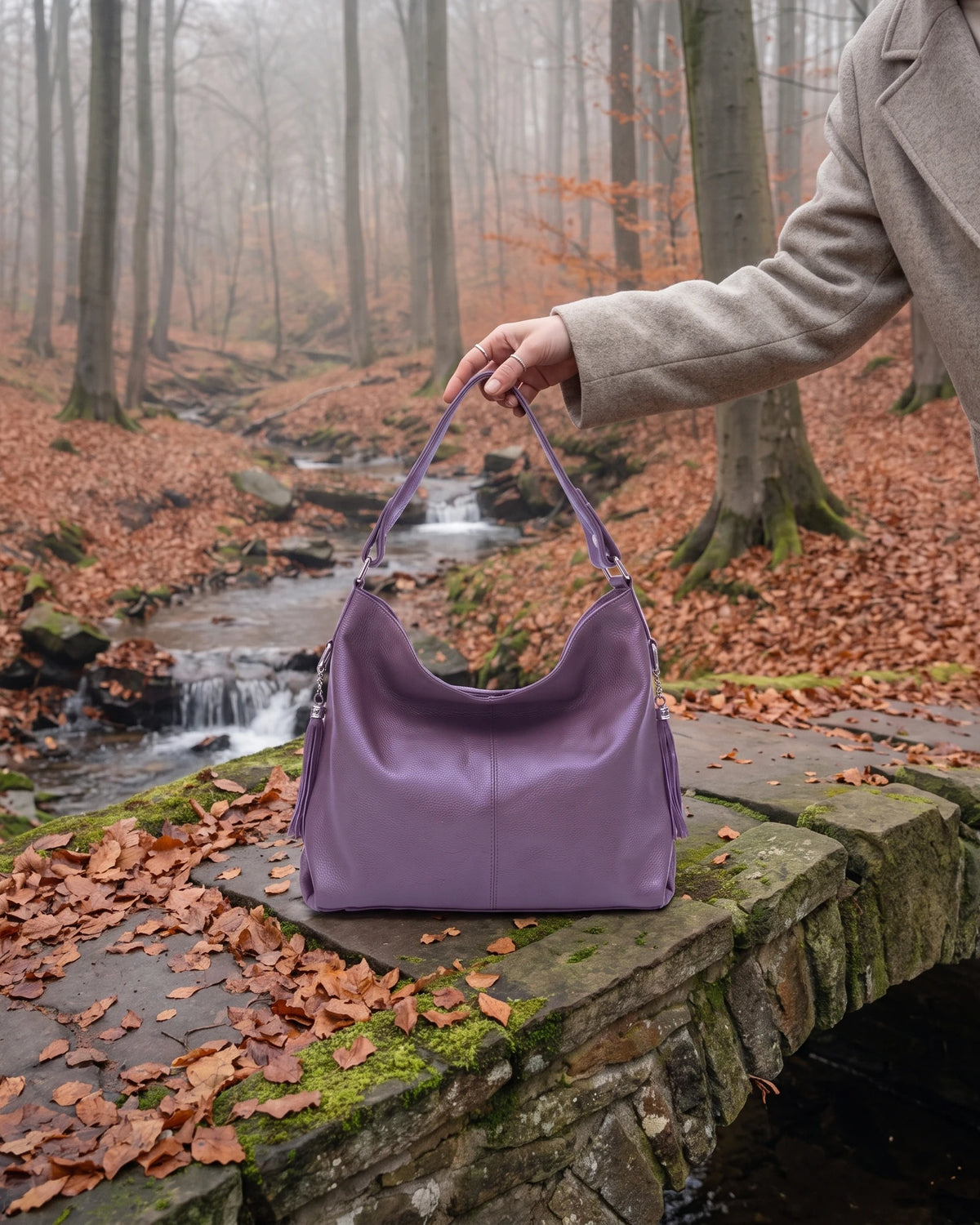 The Houle Soft in Lilas, muted lilac leather hobo bag standing on the mossy stones of a forest footbridge