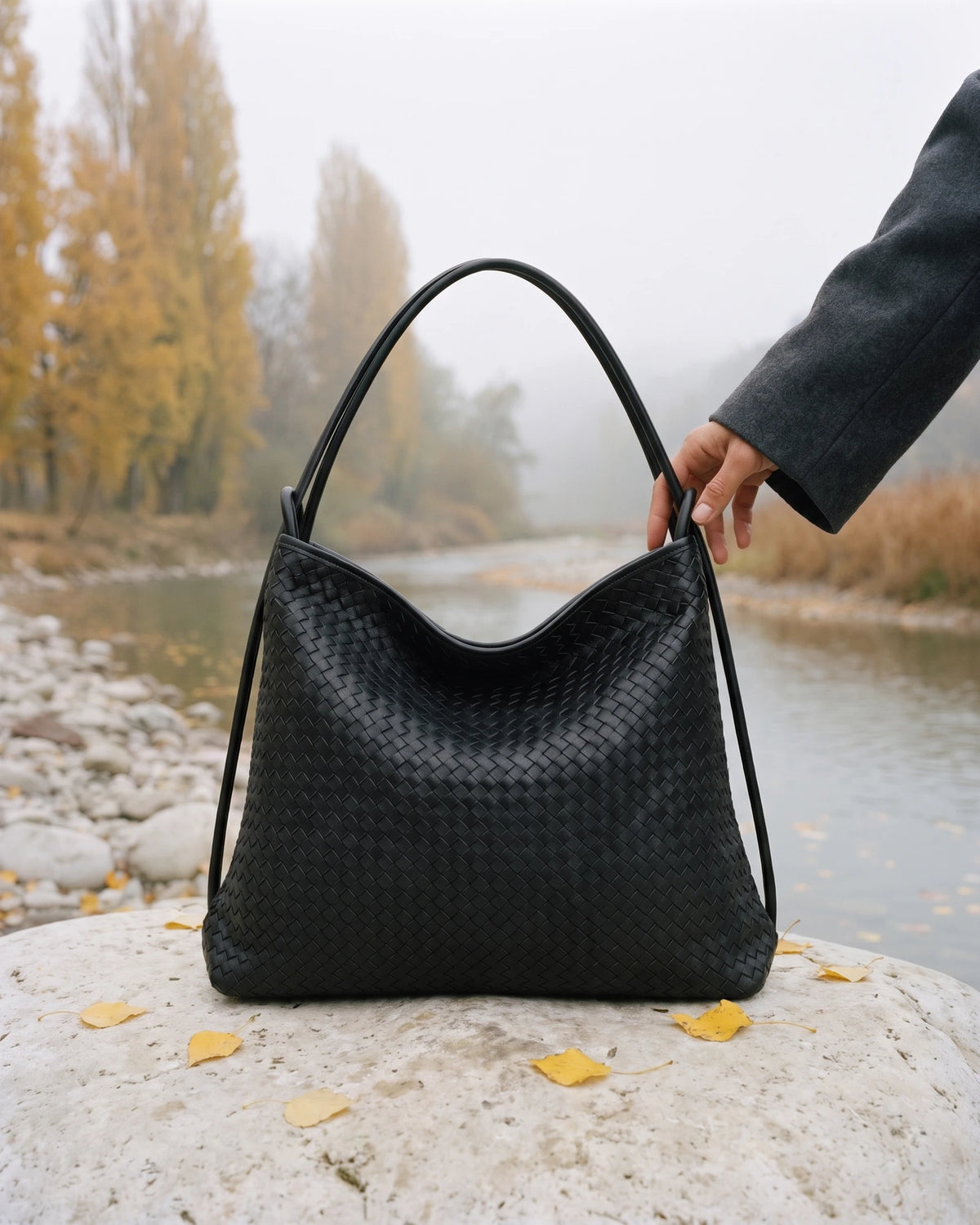 The Embellie Grand in Noir Mat, large matte black woven leather tote standing on a river boulder