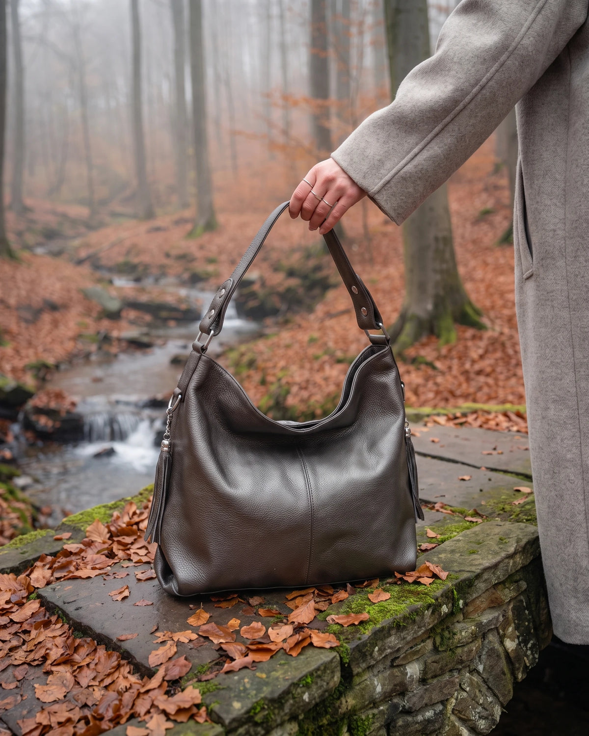 The Houle Soft in Bronze, metallic pewter leather hobo bag held on the mossy stones of a forest footbridge