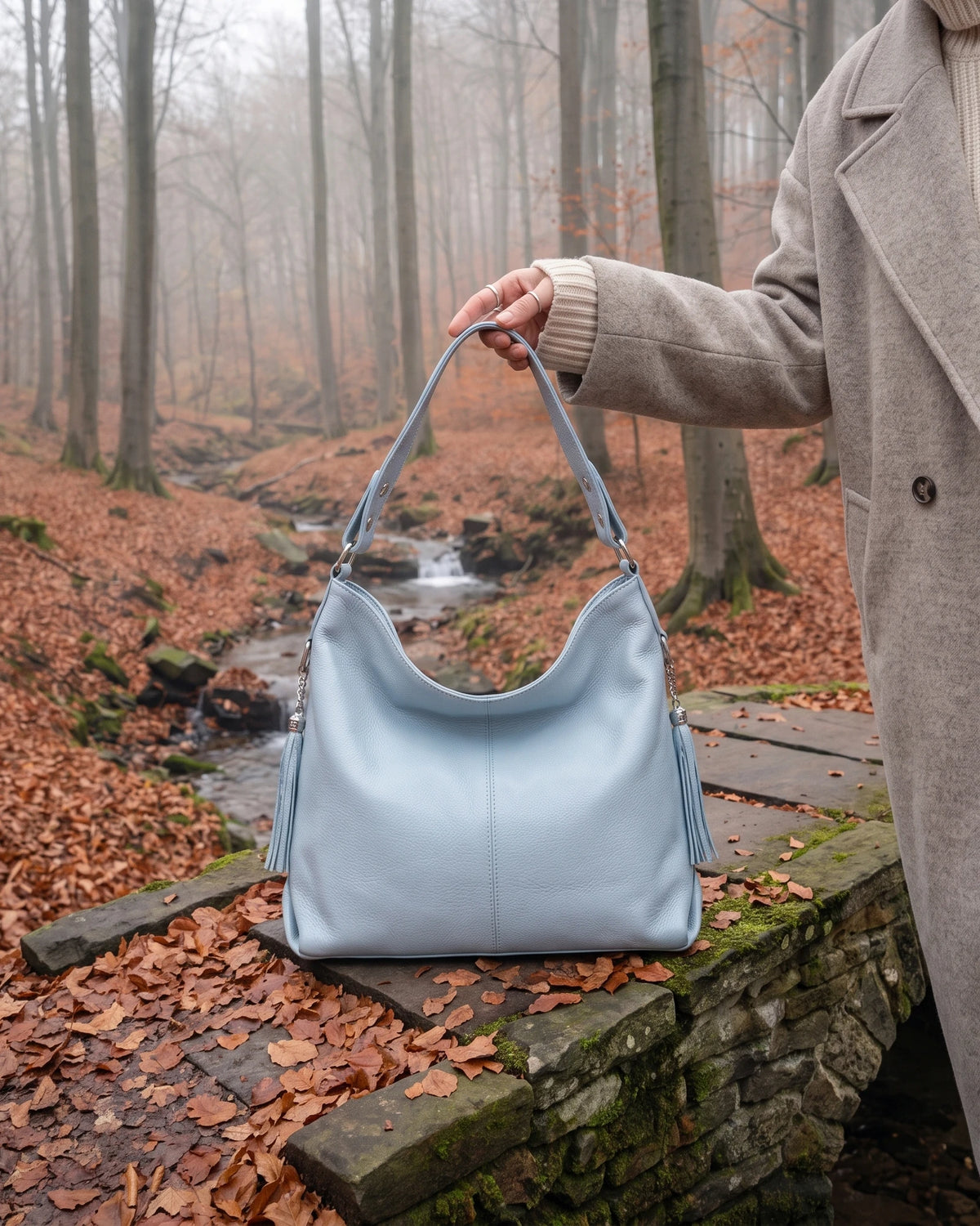 The Houle Soft in Ciel, powder blue leather hobo bag held on the mossy stones of a forest footbridge