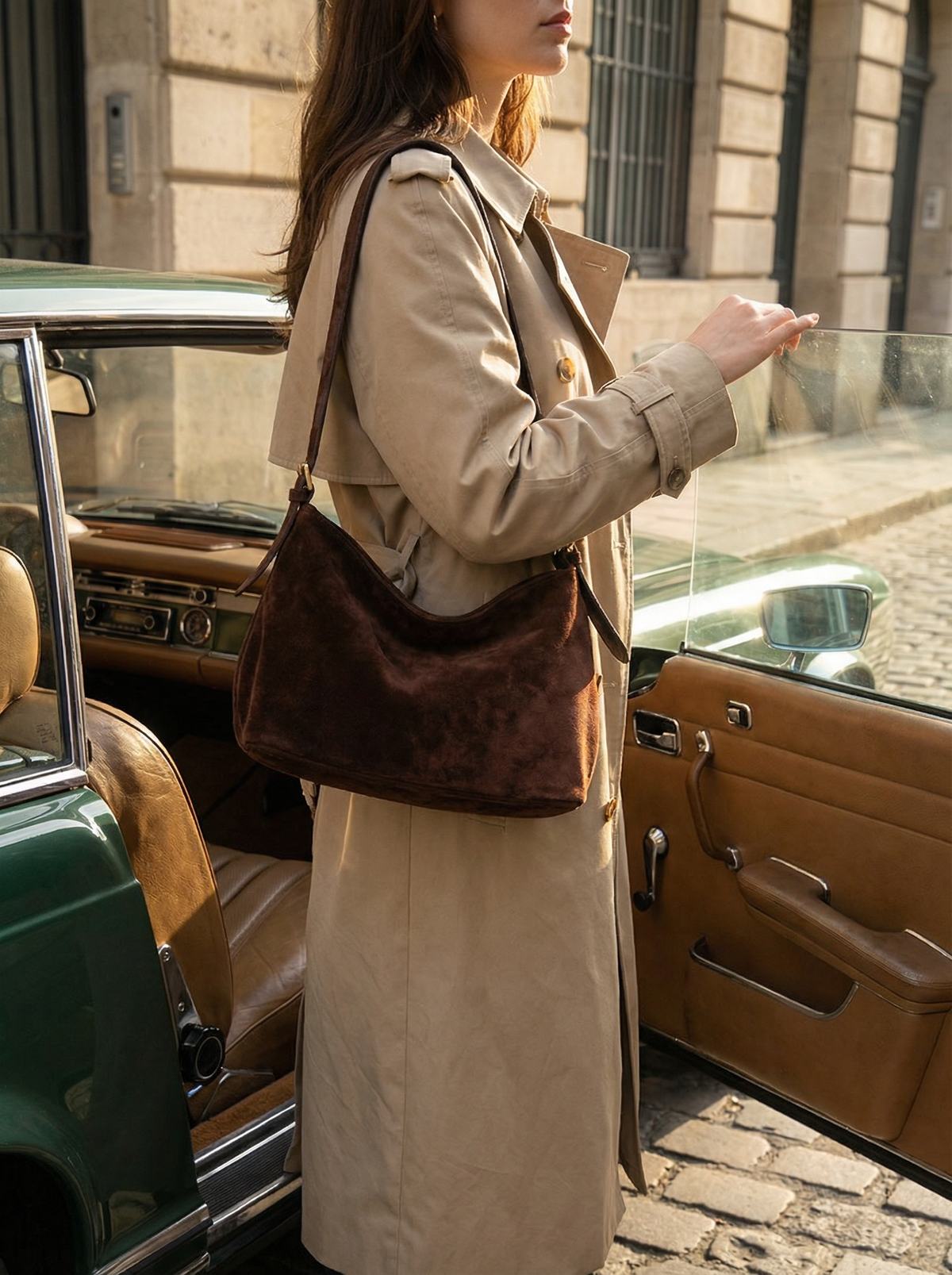 Woman in a camel trench coat carrying The Houle Petite Suede in Chocolate by a vintage car