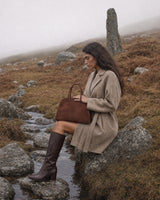 The Aurore Suede Satchel in Chocolate, carried by hand in the misty hills